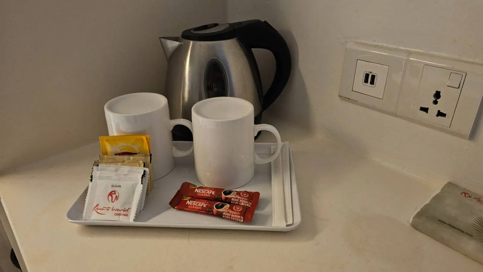 A tray on a countertop with two white mugs, an electric kettle, tea bags, coffee sachets, sugar packets, and stirrers next to a power outlet on the wall.