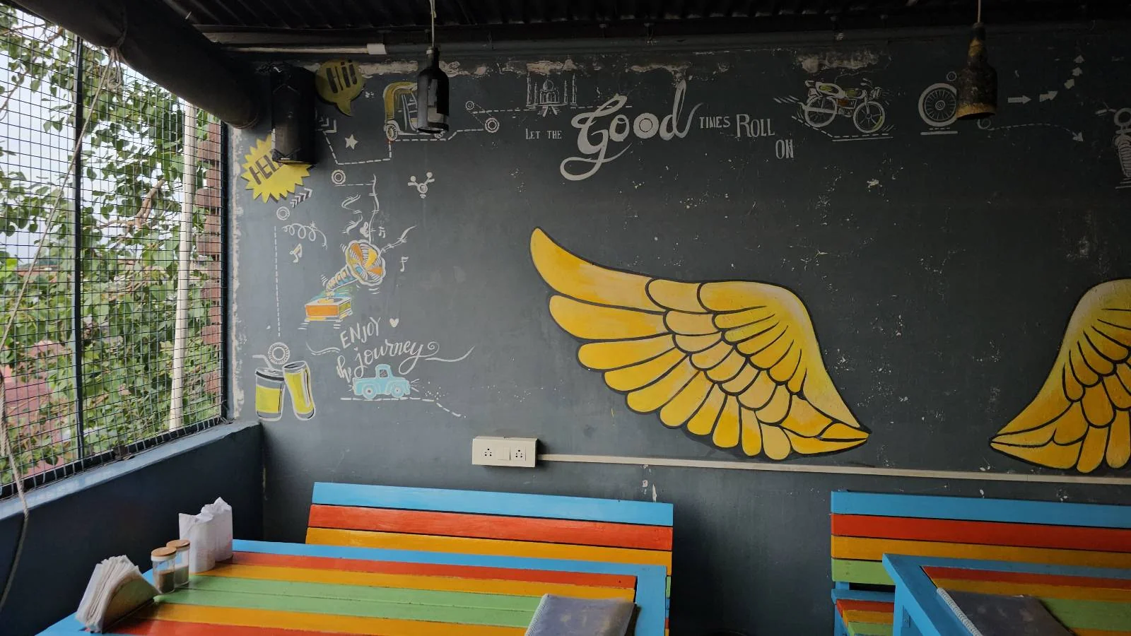 A colorful cafe interior with blue, yellow, and orange tables. The blackboard wall features painted yellow wings and doodles, including a bicycle, headphones, and the word "good." Large windows reveal greenery outside.