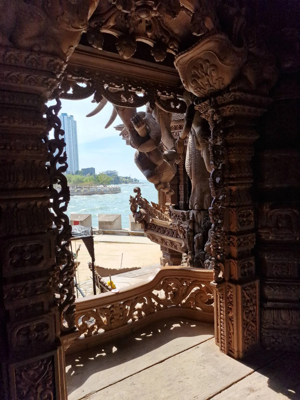 Intricately carved wooden doorway frame revealing a view of a river and a modern cityscape in the background. Sunlight highlights the detailed craftsmanship of the woodwork.