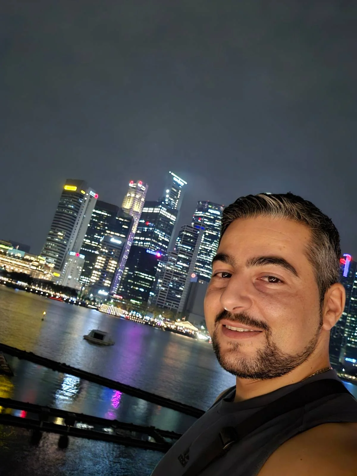 A person with short hair and a beard taking a selfie at night by a waterfront, with a brightly lit city skyline in the background. The buildings and their reflections create a colorful scene against the dark sky.