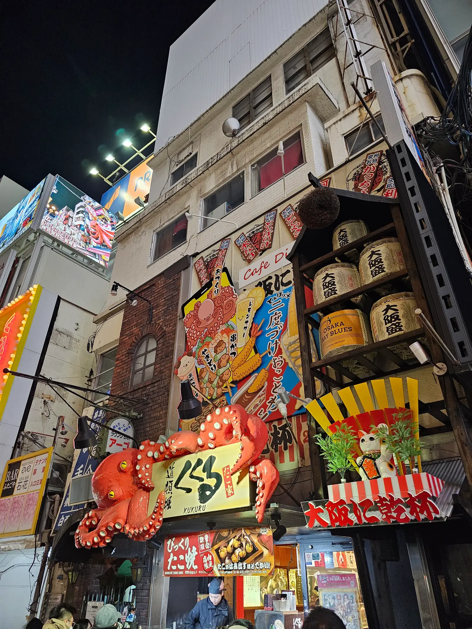 Colorful building facade at night, decorated with large lanterns, a red crab sculpture, and vibrant murals. People stand near the entrance, and bright lights illuminate the lively street scene.