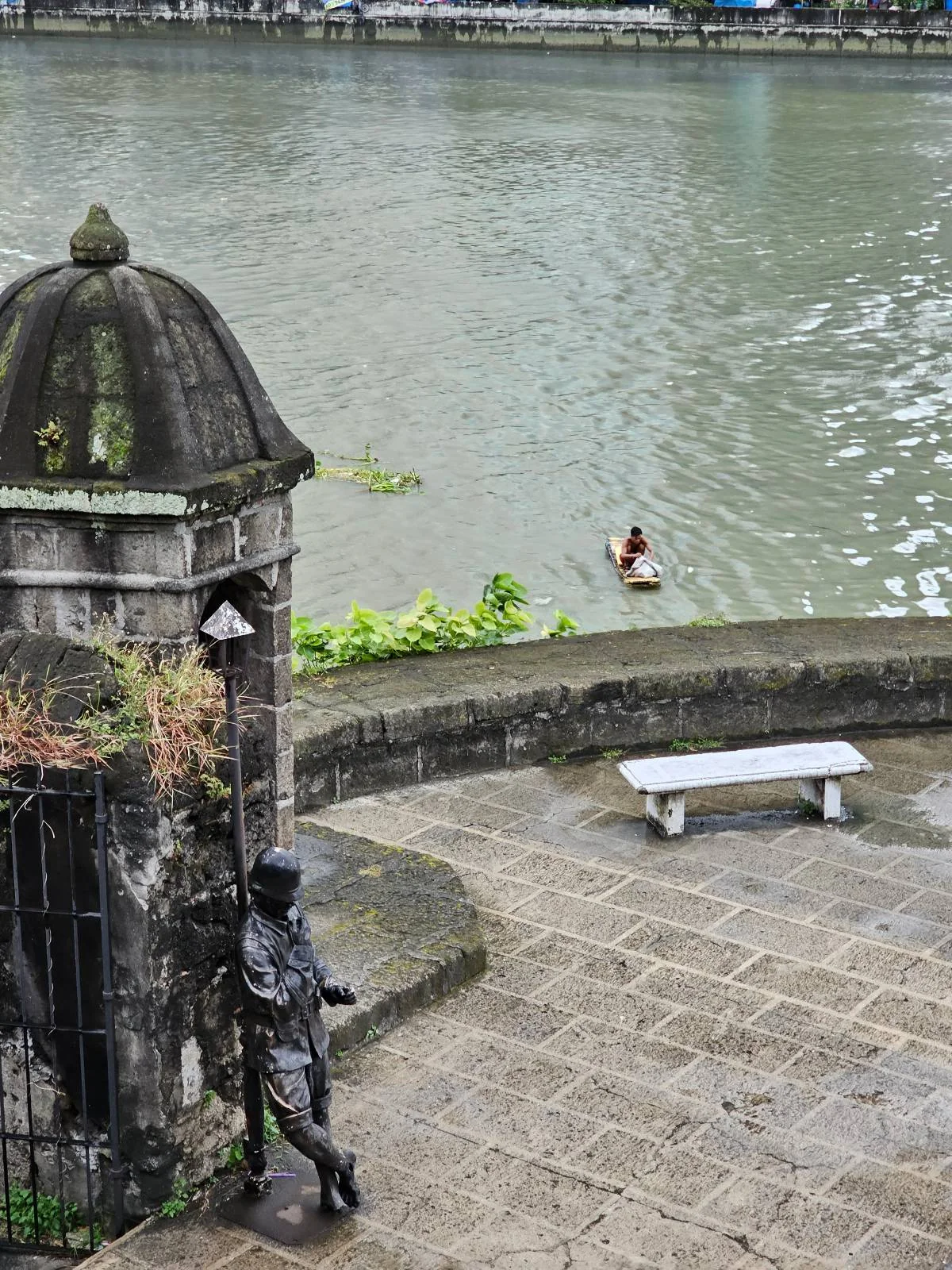 A stone sculpture of a person leaning against a small, domed structure by a riverside. A lone duck floats in the river. A stone bench sits on the paved walkway beside the river. Overcast sky reflects on the water.