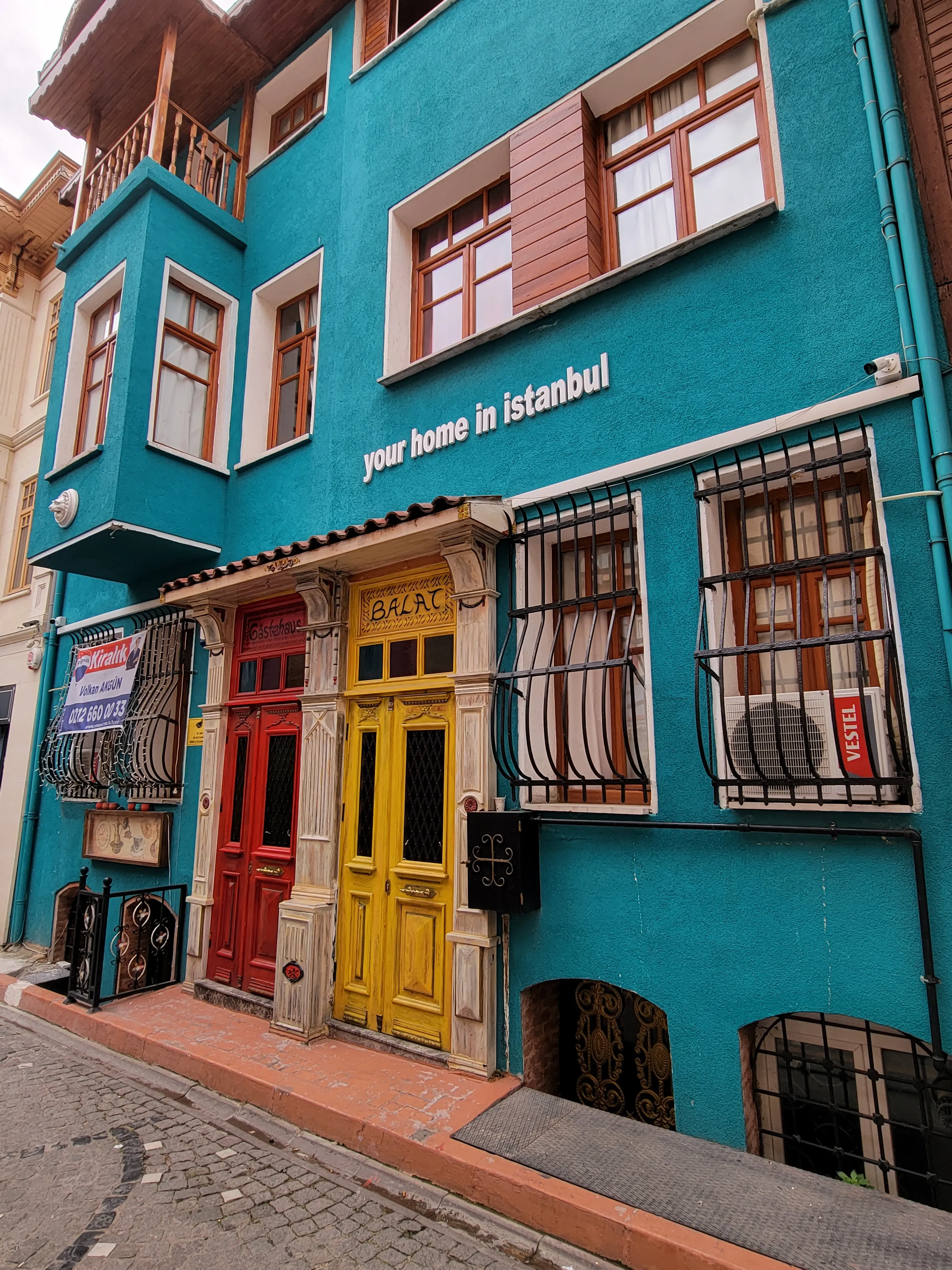 A vibrant blue building with yellow and red doors, barred windows, and a sign reading "Your Home in Istanbul" on a sloped street in a city.