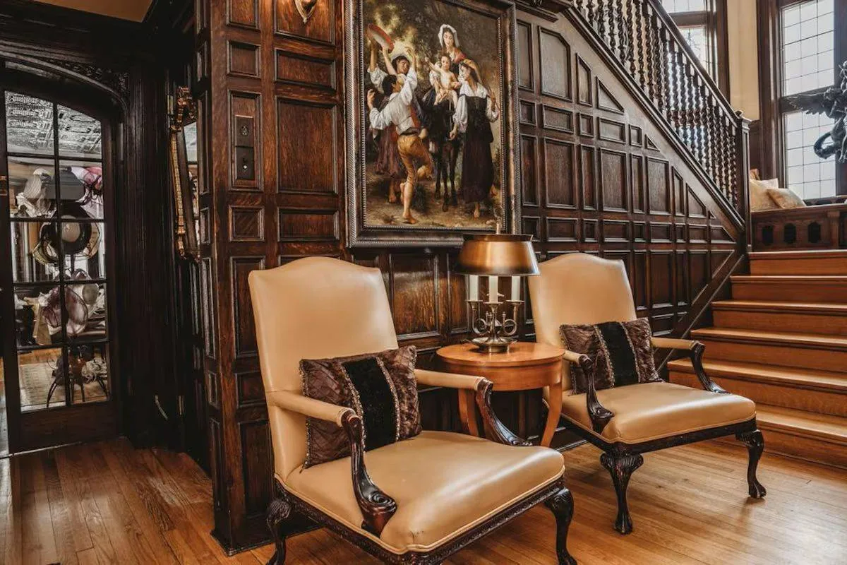 Two beige armchairs with cushions beside a wooden staircase. A round table with a lamp is between them. A framed painting on the wall. Classic decor.