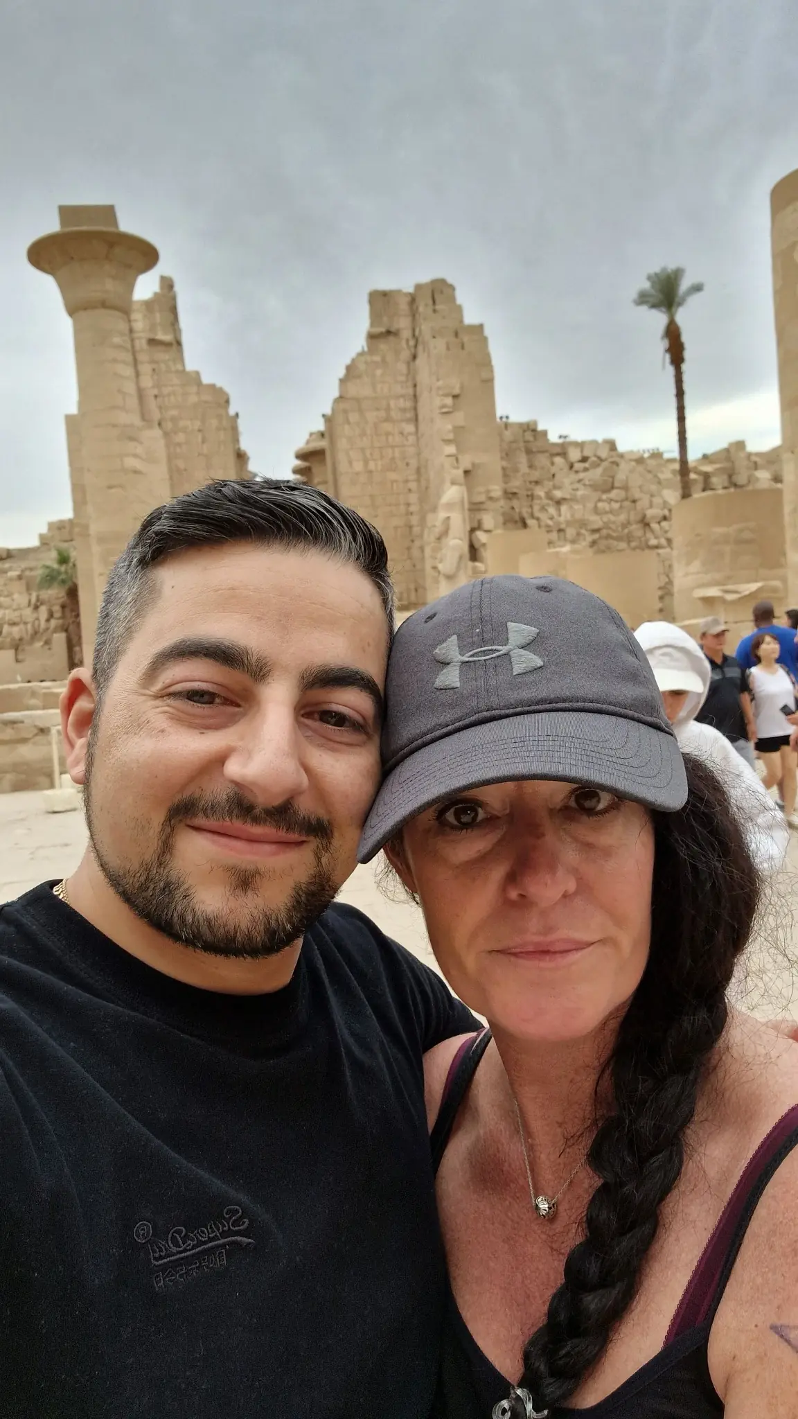 Two people pose for a selfie in front of an ancient stone structure, with other tourists and palm trees visible in the background.