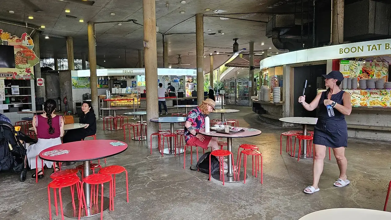 An open-air food court with red stools and circular tables. People are sitting and enjoying their meals. One person is walking while holding a phone. Various food stalls are visible in the background.