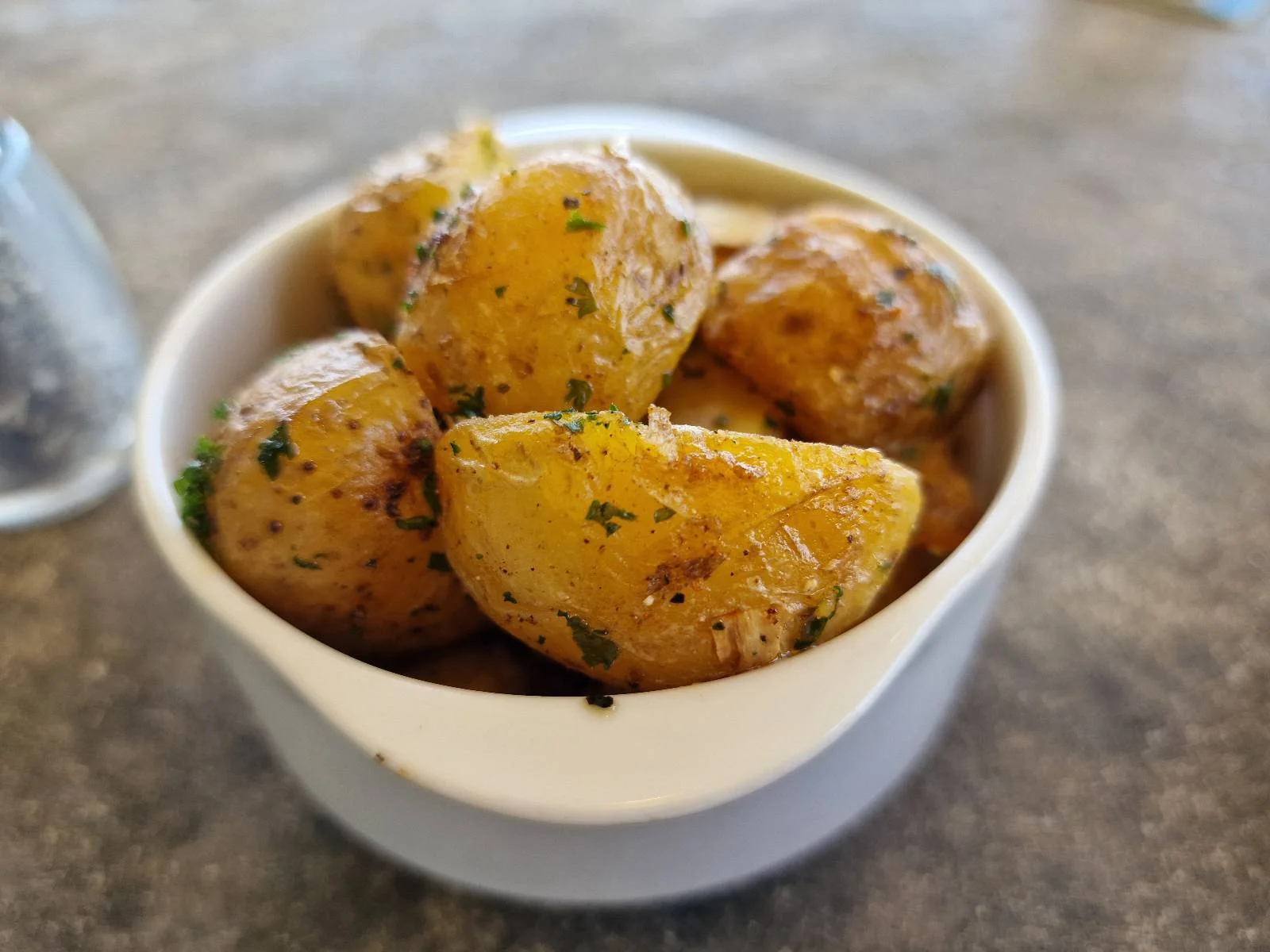 A white bowl filled with seasoned baby potatoes, garnished with chopped herbs. The potatoes have a glossy, lightly peppered appearance, placed on a gray textured surface.