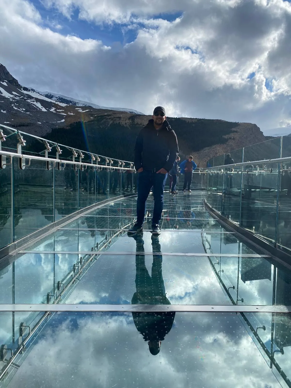 A person stands on a glass bridge with mountains and cloudy sky in the background. The glass surface reflects the clouds and the person's silhouette.