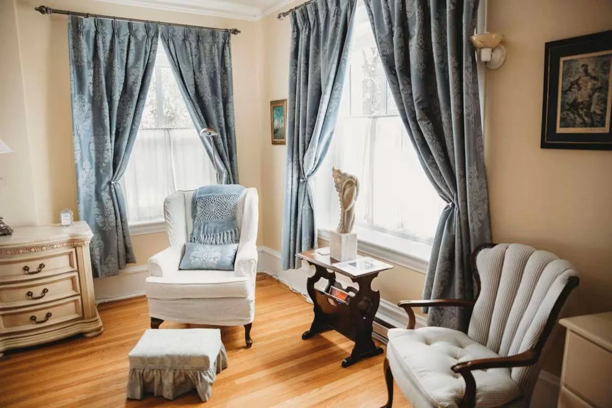 Cozy living room with blue patterned curtains, white armchair, and a small table with a sculpture. Warm light and a calm atmosphere.