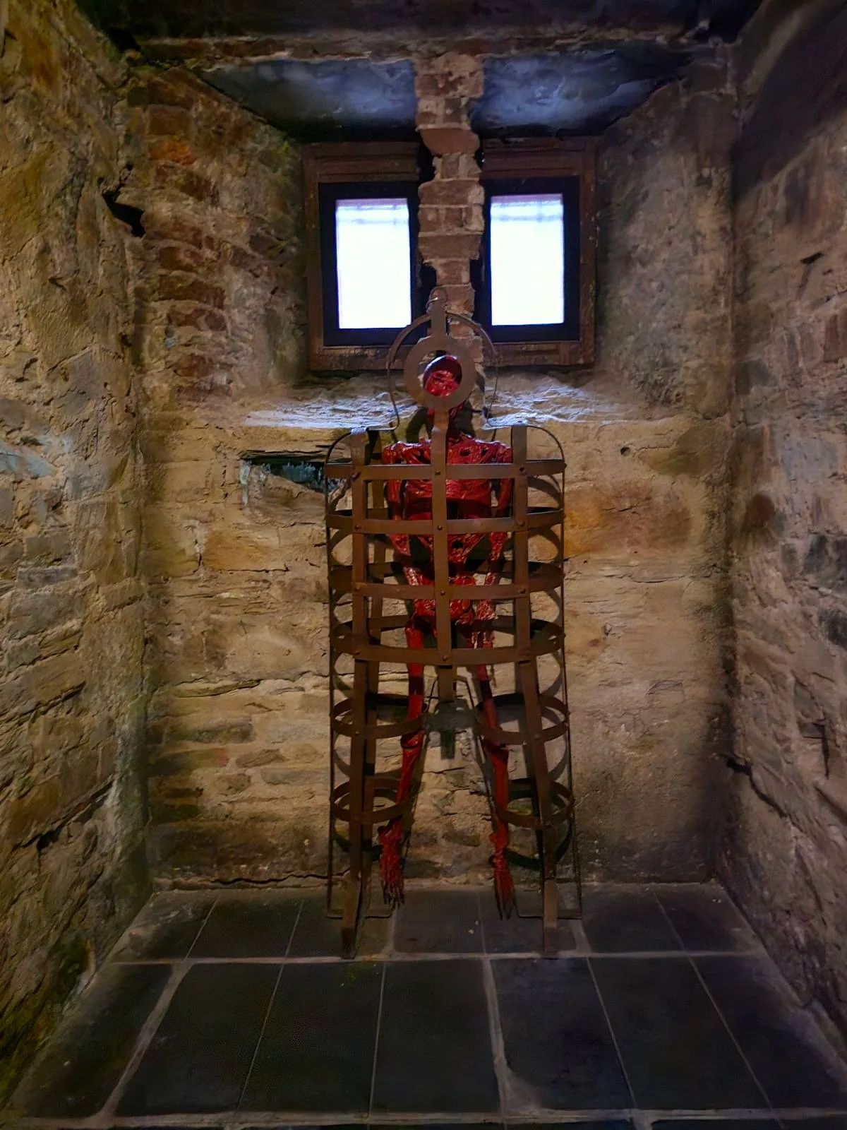 A medieval wooden torture device, resembling a rack, stands in a small stone room with two small windows illuminated by soft light.