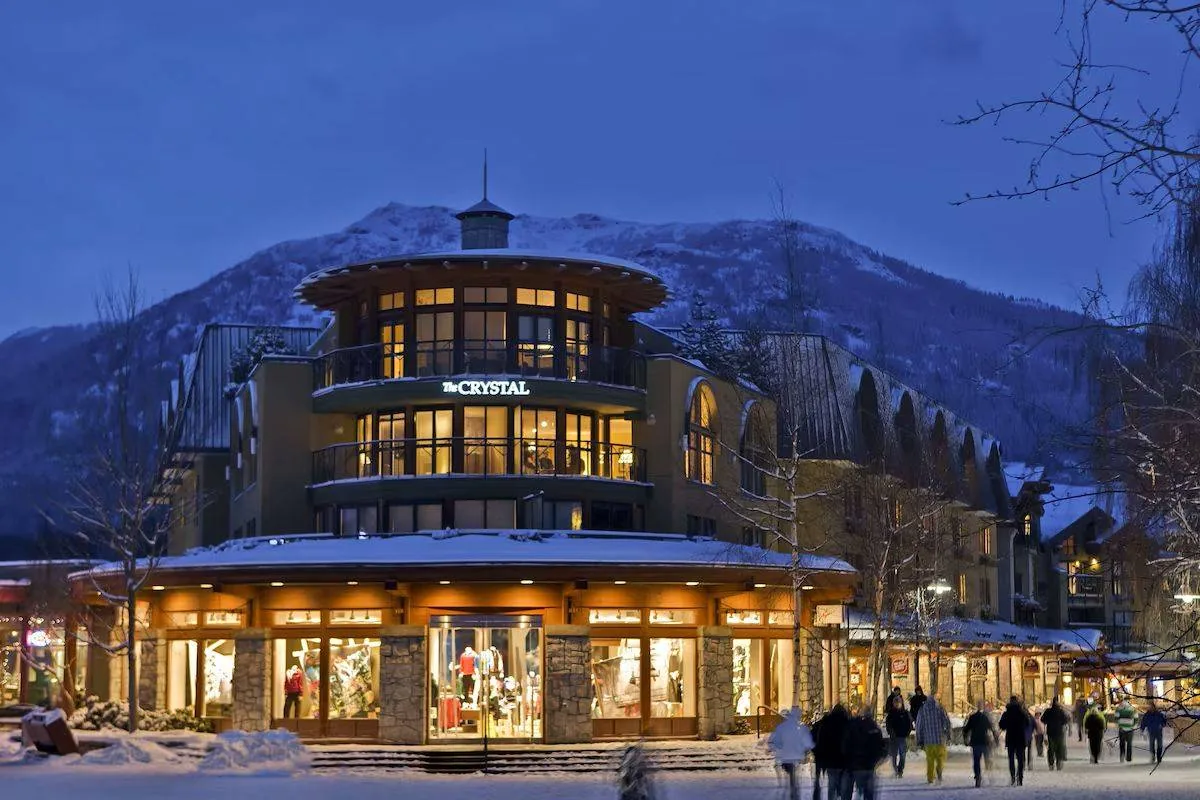 A bustling shopping area at dusk with people walking past a circular building labeled "Mountain." The building displays brightly lit storefronts. Snow-covered mountains are visible in the background under a deep blue sky.