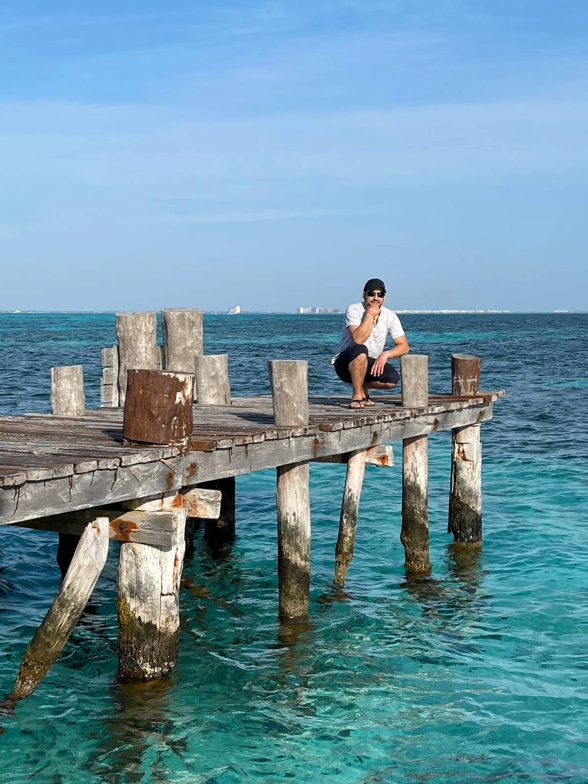 A person crouches on the edge of an old wooden pier jutting out into clear turquoise water, under a bright blue sky. The sea is calm, and distant ships are visible on the horizon.