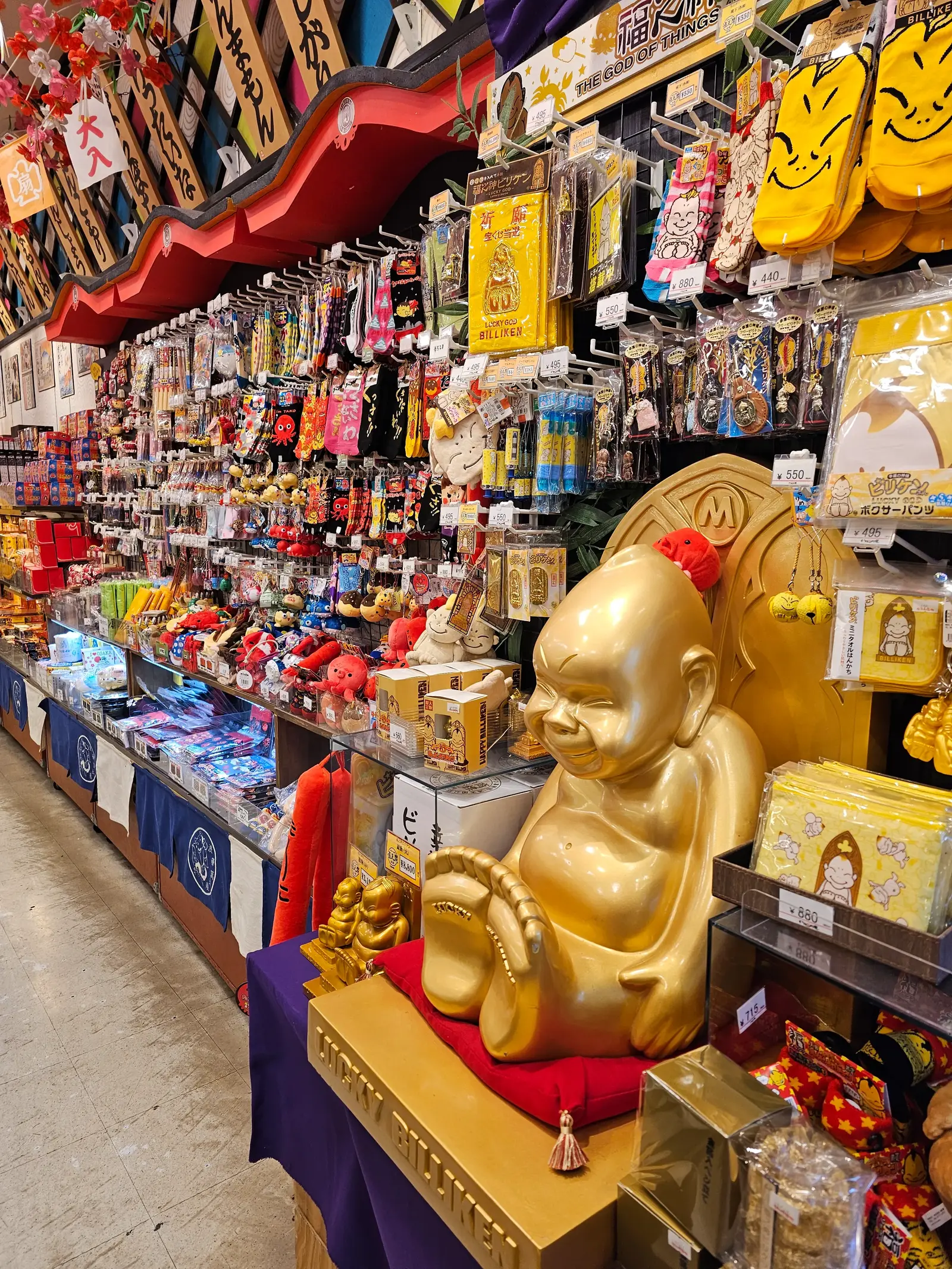 A colorful store display features a large golden Buddha statue in the foreground, surrounded by shelves filled with various Asian trinkets, souvenirs, and decorative items.