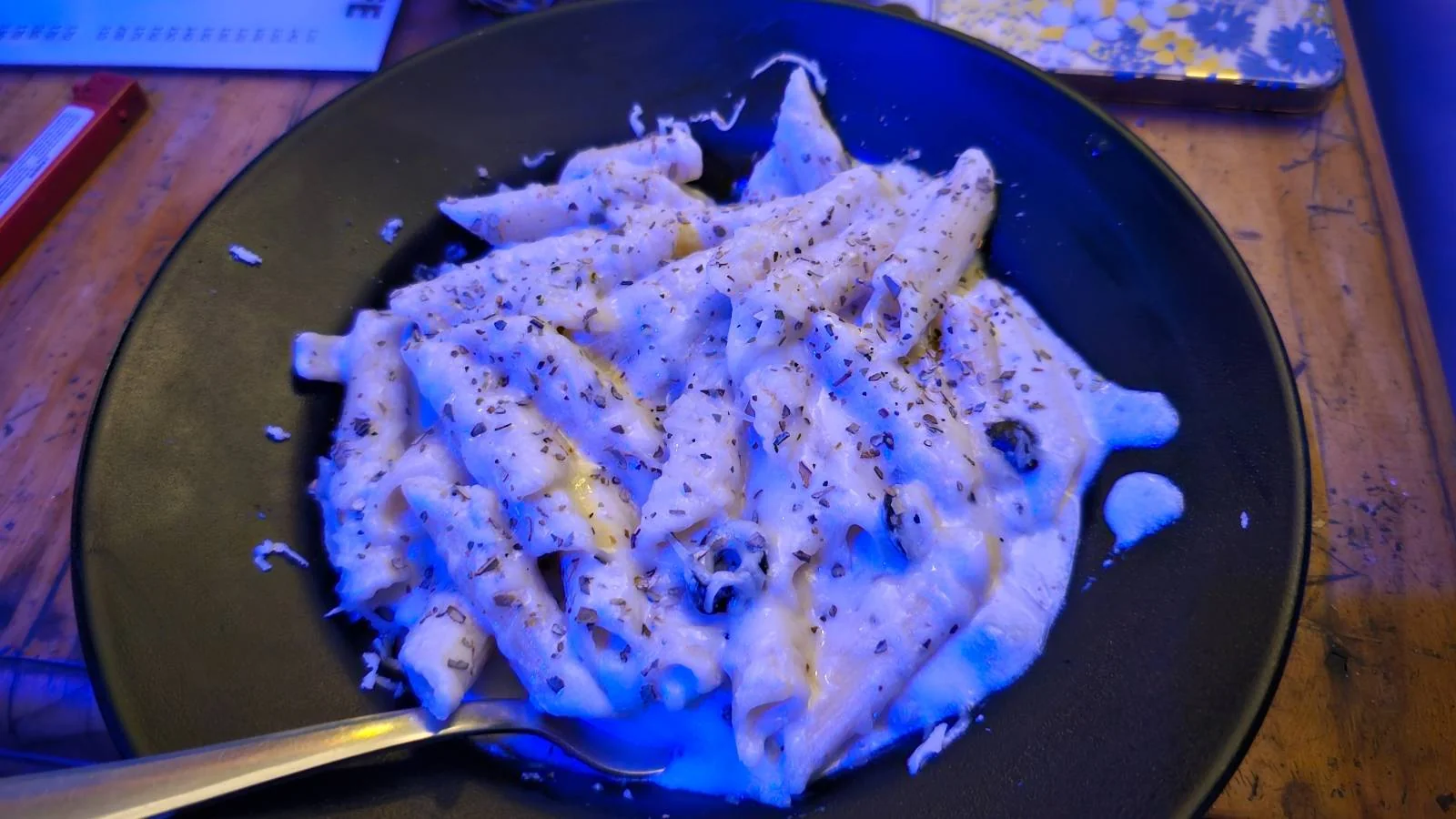 A black plate filled with penne pasta covered in creamy white sauce, topped with black pepper and grated cheese, sits on a wooden table.