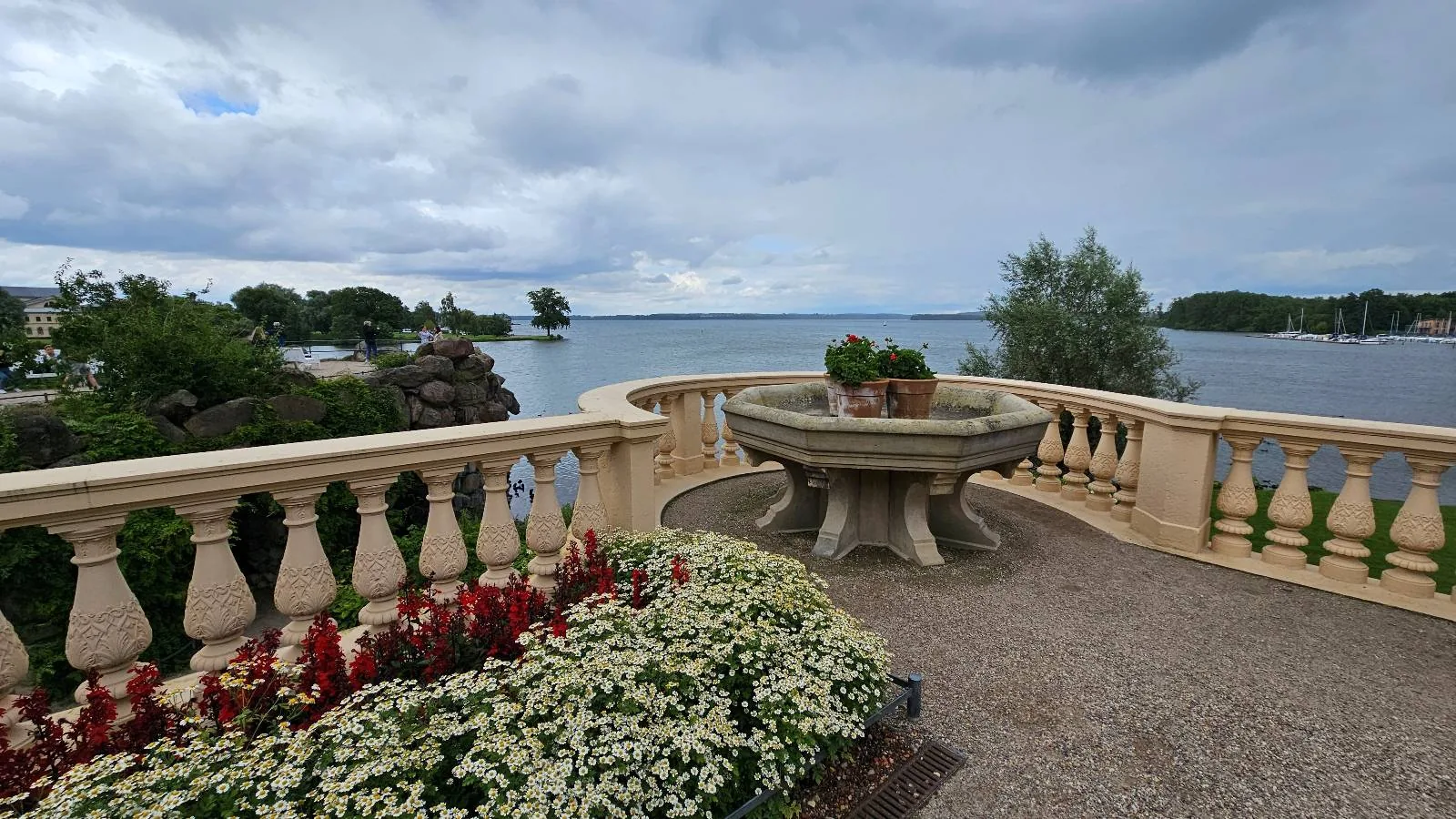 A stone terrace with a large round planter overlooks a calm lake, surrounded by green trees and cloudy skies. Red and white flowers bloom along the railing in the foreground.