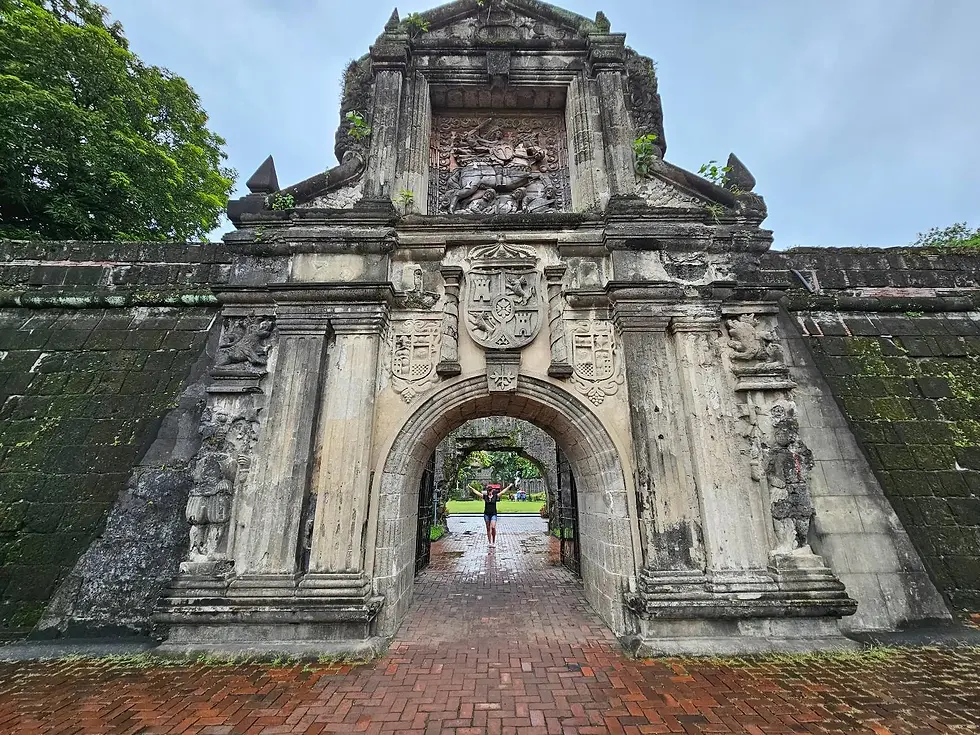 Stone archway at Fort Santiago with detailed carvings. A person walks beneath, set against a cloudy sky and lush greenery.