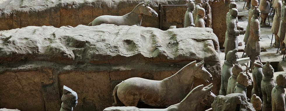 Rows of life-sized terracotta horses and soldiers stand in an ancient excavation site, partially surrounded by earth and clay walls, representing China’s famous Terracotta Army.