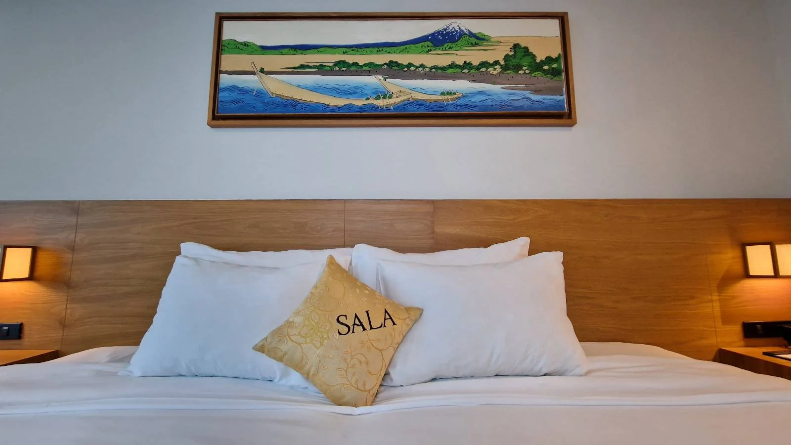 A neatly made bed with white linens and a beige pillow with "SALA" on it. The headboard is wooden, and two warm-colored lamps are mounted on each side. Above the bed is a landscape painting of a mountain and water scene.