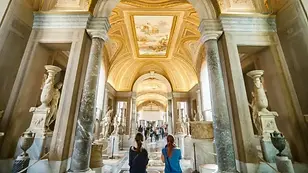 Visitors walking through an ornate gallery inside the Vatican Museums with marble statues and frescoed ceilings, Rome, Italy.