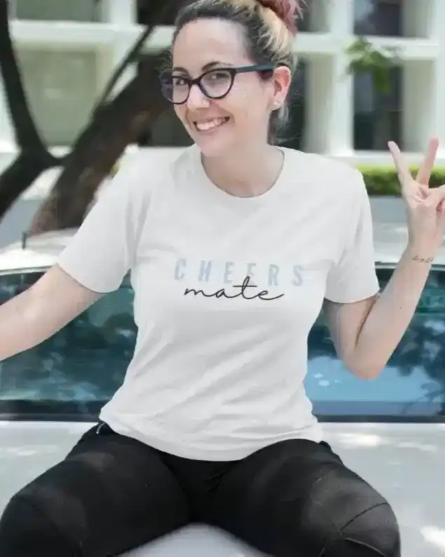 A person wearing glasses and a white t-shirt with the text "CHEERS mate" sits on a car, smiling and making a peace sign.