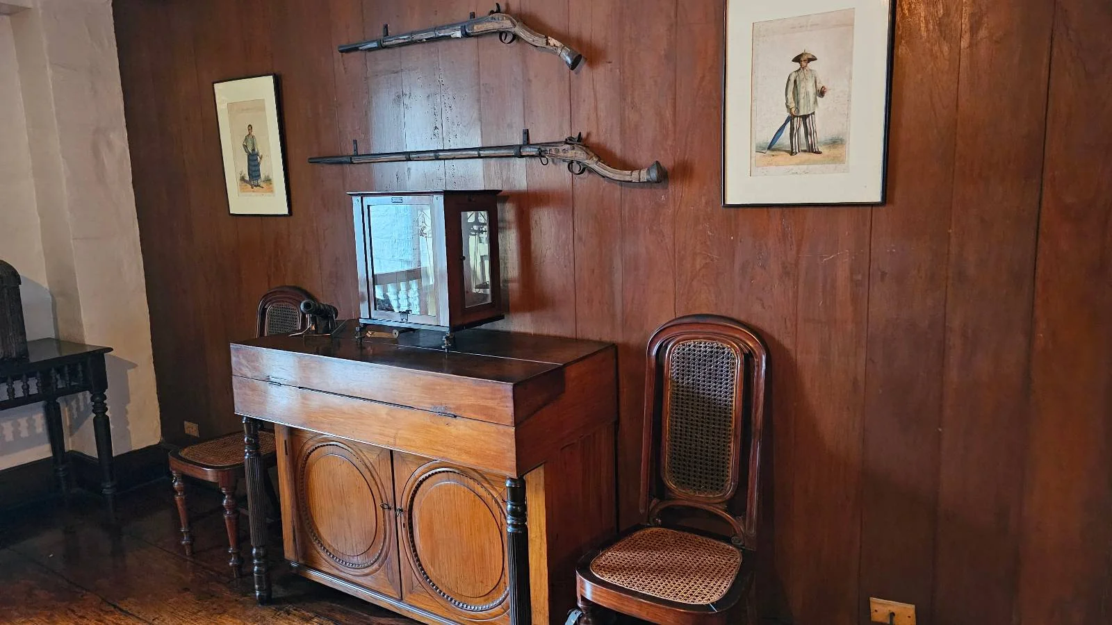 A vintage room with wooden walls features two rifles mounted horizontally, a cabinet with glass doors, and two framed pictures. A wooden chair with a woven seat is positioned next to the cabinet. The floor is dark wood.