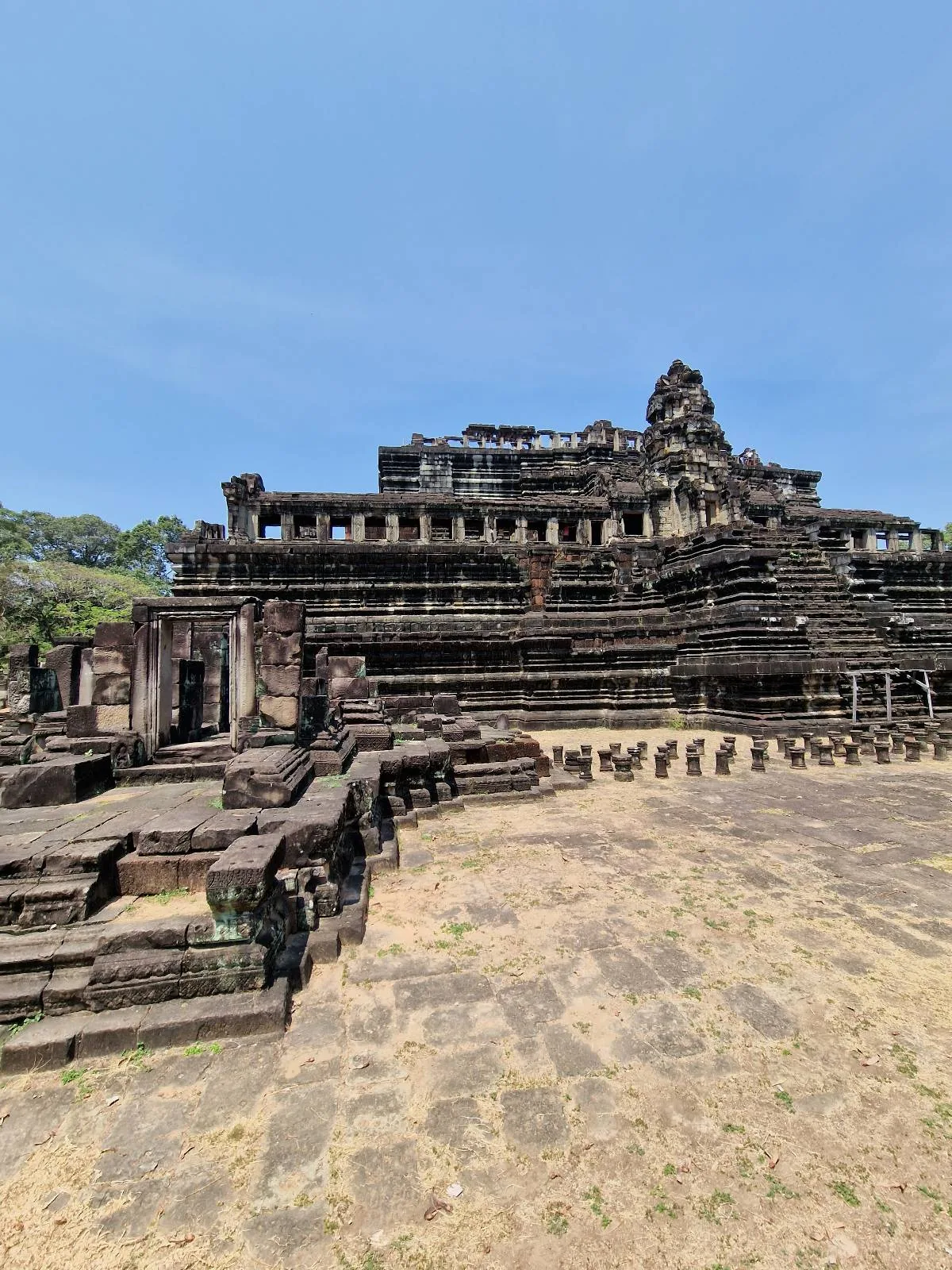 Ancient stone temple with multiple stepped terraces and intricate carvings, surrounded by greenery under a clear blue sky. The structure appears weathered and historic.