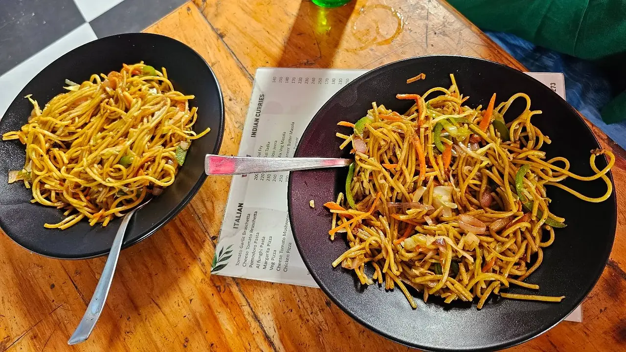 Two black bowls filled with stir-fried noodles, vegetables, and shredded meat sit on a wooden table. One bowl is larger than the other and a menu and utensils are visible between them.