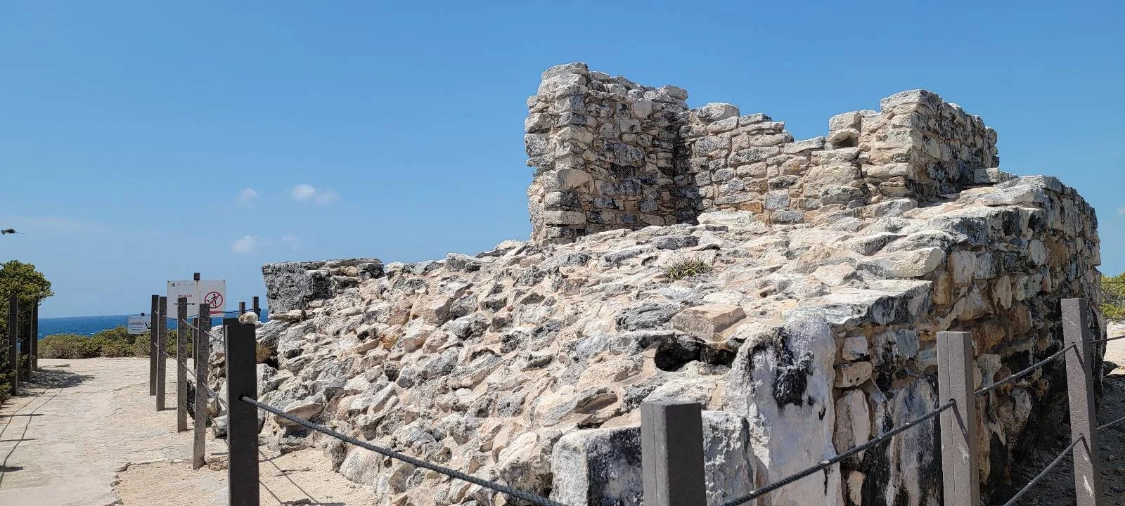 A weathered stone ruin with irregular rocks stacked to form partial walls stands under a clear blue sky. Surrounding the structure, metal posts with ropes create a barrier, and the distant blue ocean is visible to the left.