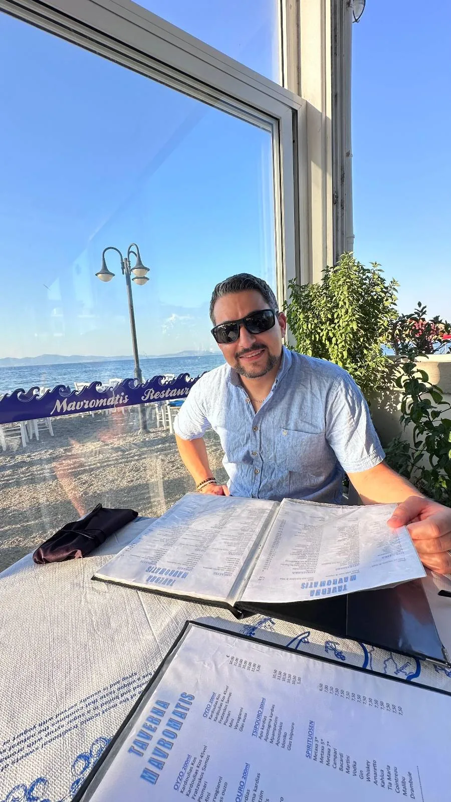 A man wearing sunglasses and a light blue shirt sits at an outdoor restaurant table with menus, seaside umbrellas, and the ocean visible through large windows under a clear blue sky.