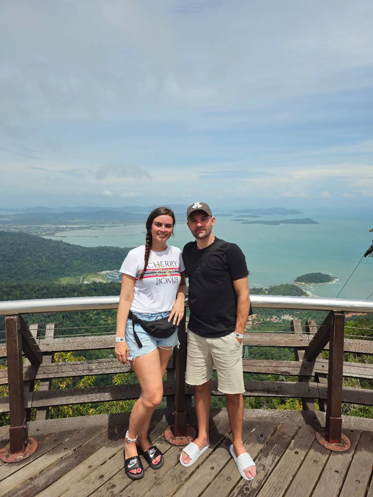 A woman and a man stand on a wooden viewing platform overlooking a scenic landscape with blue waters and green hills. The sky is partly cloudy. The woman wears a white shirt and shorts, while the man wears a black t-shirt and shorts.