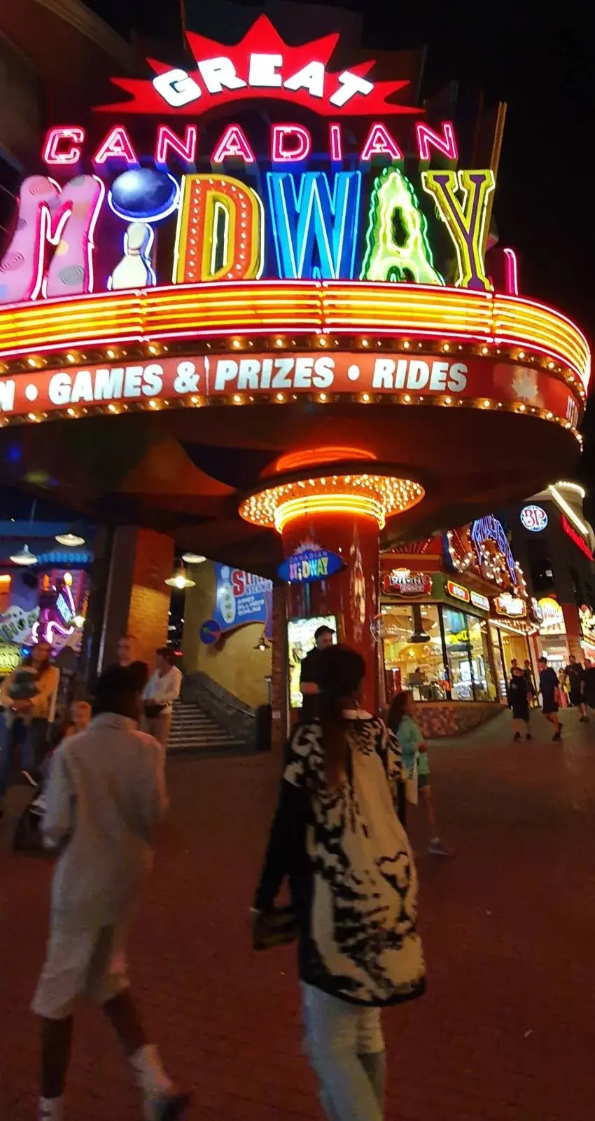 Colorful neon sign for "Great Canadian Midway" at night. People walk below, creating a lively, fun atmosphere.