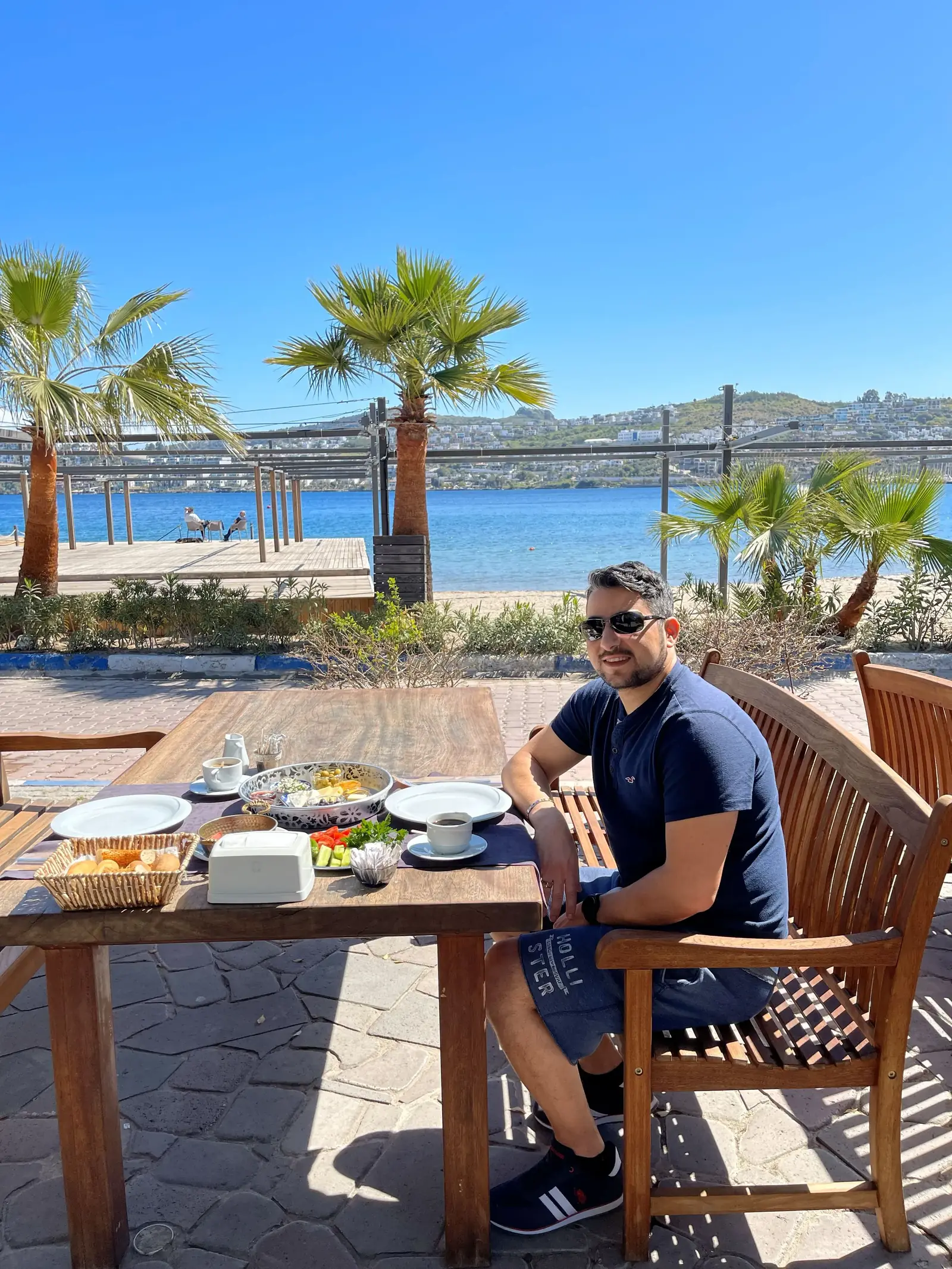A man wearing sunglasses sits at an outdoor table set for a meal, with plates of food in front of him. Palm trees, a sandy beach, and a calm body of water are visible in the sunny background.
