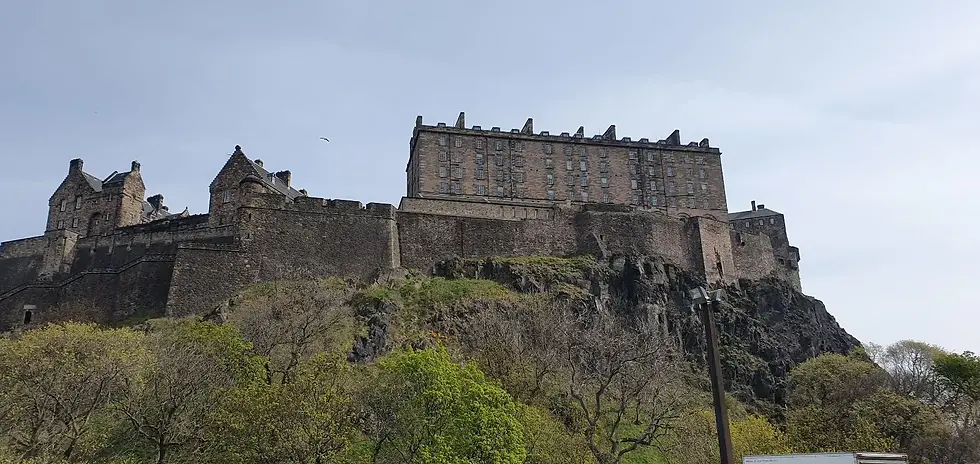 Edinburgh Castle on a rocky hill above trees under a pale blue sky, with stone fortress walls and towers.