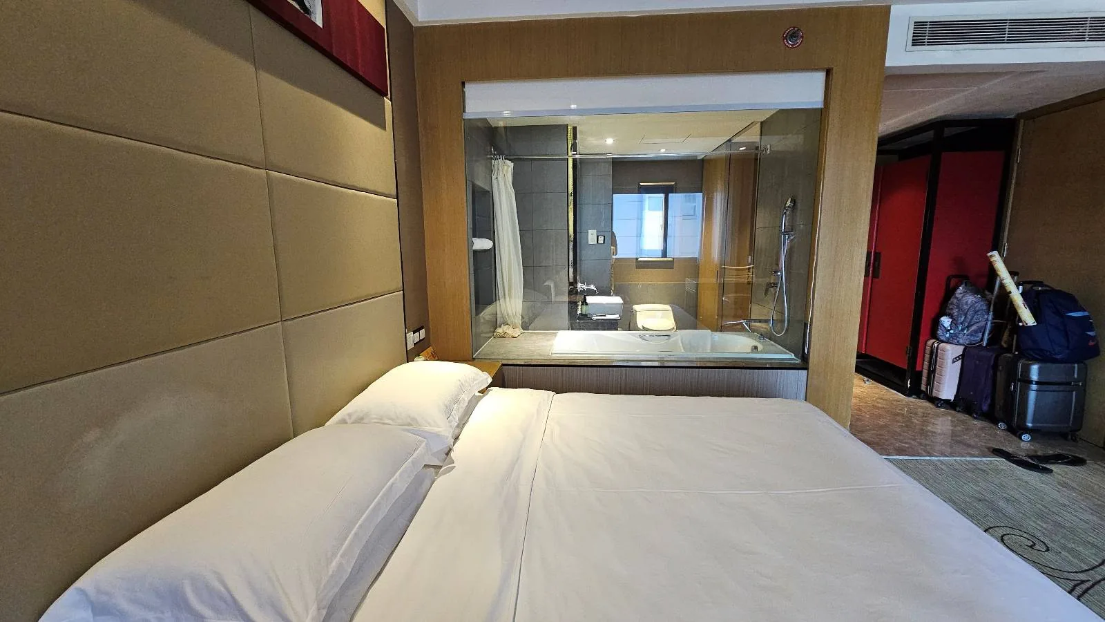 A modern hotel room with a neatly made bed in the foreground; behind the bed is a glass wall revealing a bathroom with a shower and bathtub, and luggage is visible near the entrance.