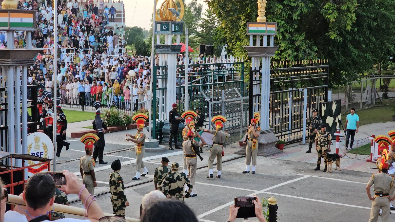 A crowd watches as Indian and Pakistani soldiers in ceremonial uniforms perform the Wagah border ceremony at an open gate, with onlookers taking photos and standing behind fences.