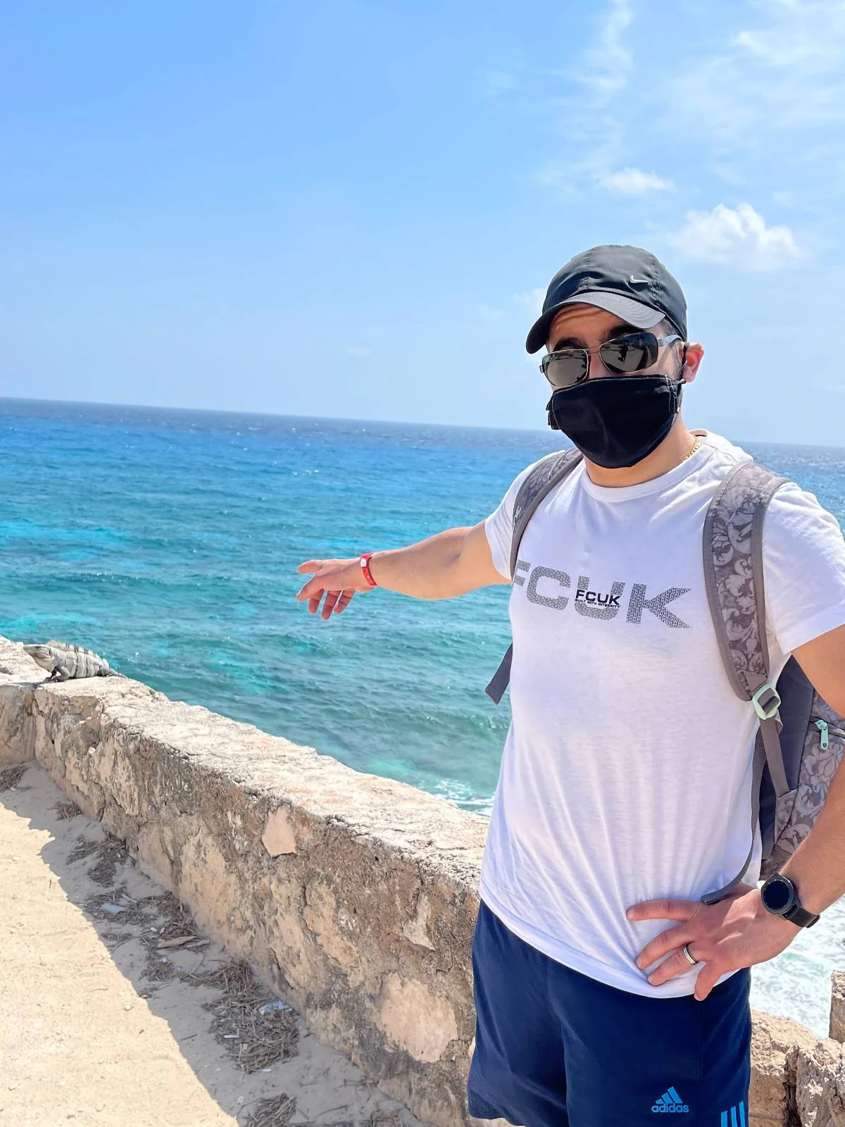 A person wearing a black mask, sunglasses, a cap, and a backpack stands near a stone wall by the ocean. They are pointing towards the blue water under a clear sky.