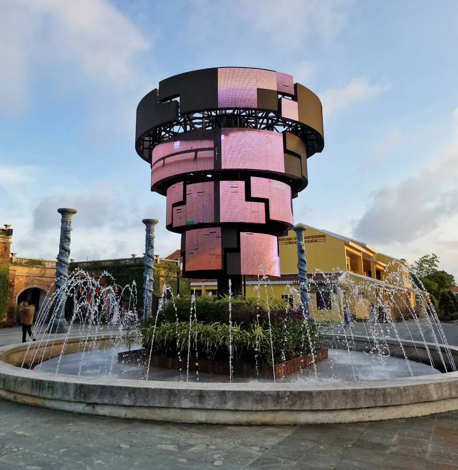 Modern cylindrical fountain with digital led screens, surrounded by jets of water and plants, under a partly cloudy sky.