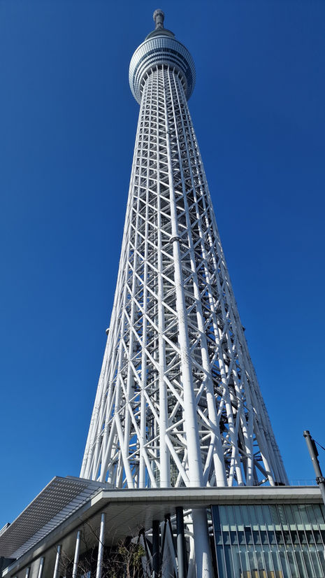 The image captures Tokyo SkyTree, the tallest tower with its iconic tall, white lattice structure set against a clear blue sky. The cylindrical top section of this third highest free-standing structure rises gracefully above modern buildings at its base.