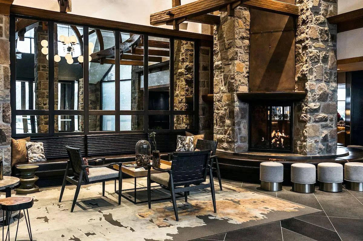 Modern lodge interior with stone fireplace, wooden beams, and large windows. Black chairs and a wooden table are on a cowhide rug. Circular stools line the hearth, and pendant lights hang from the ceiling, creating a cozy atmosphere.