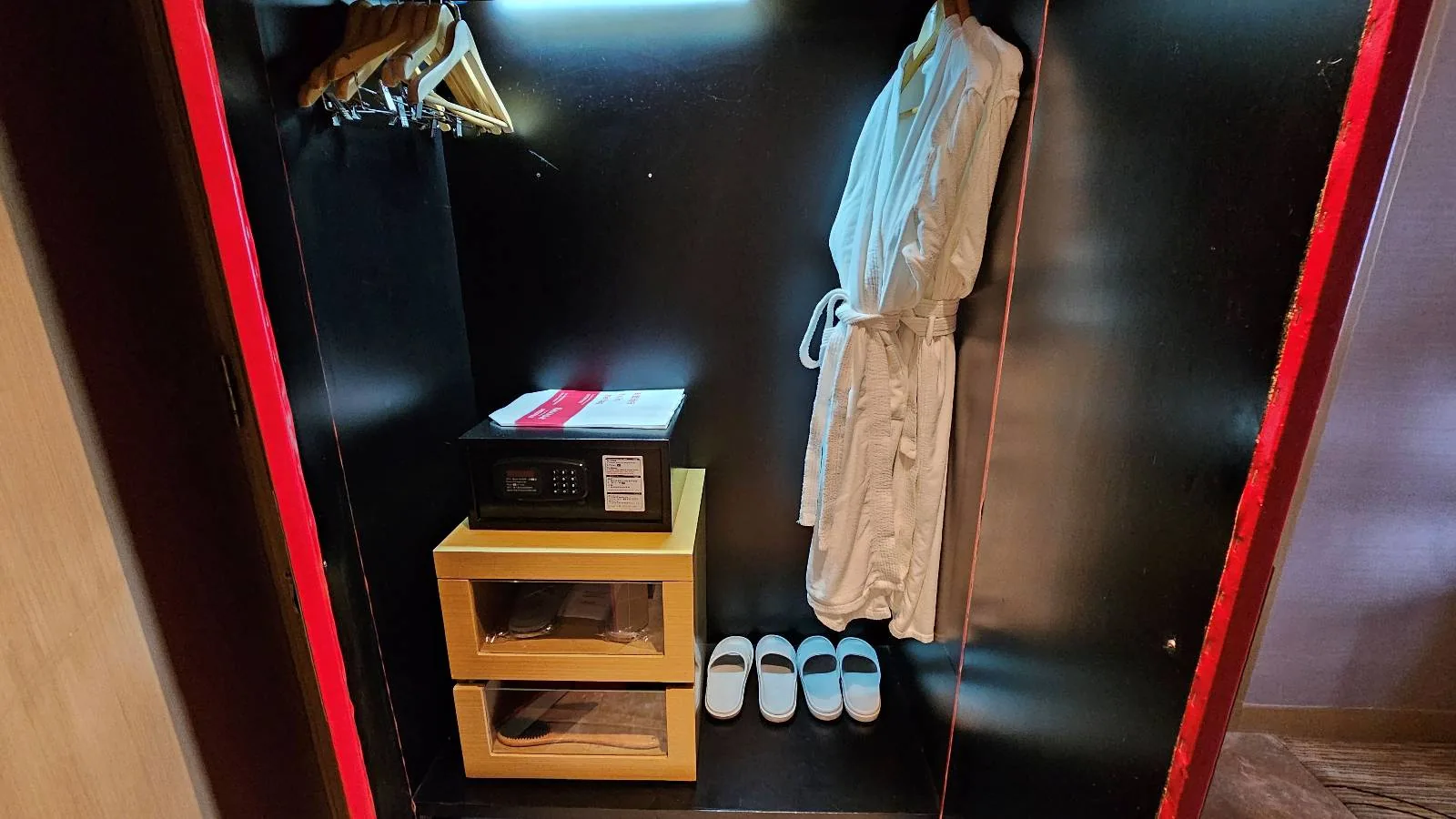 An open wardrobe with hangers, a white bathrobe, a safe, two drawers, and three pairs of slippers neatly arranged on the bottom shelf.