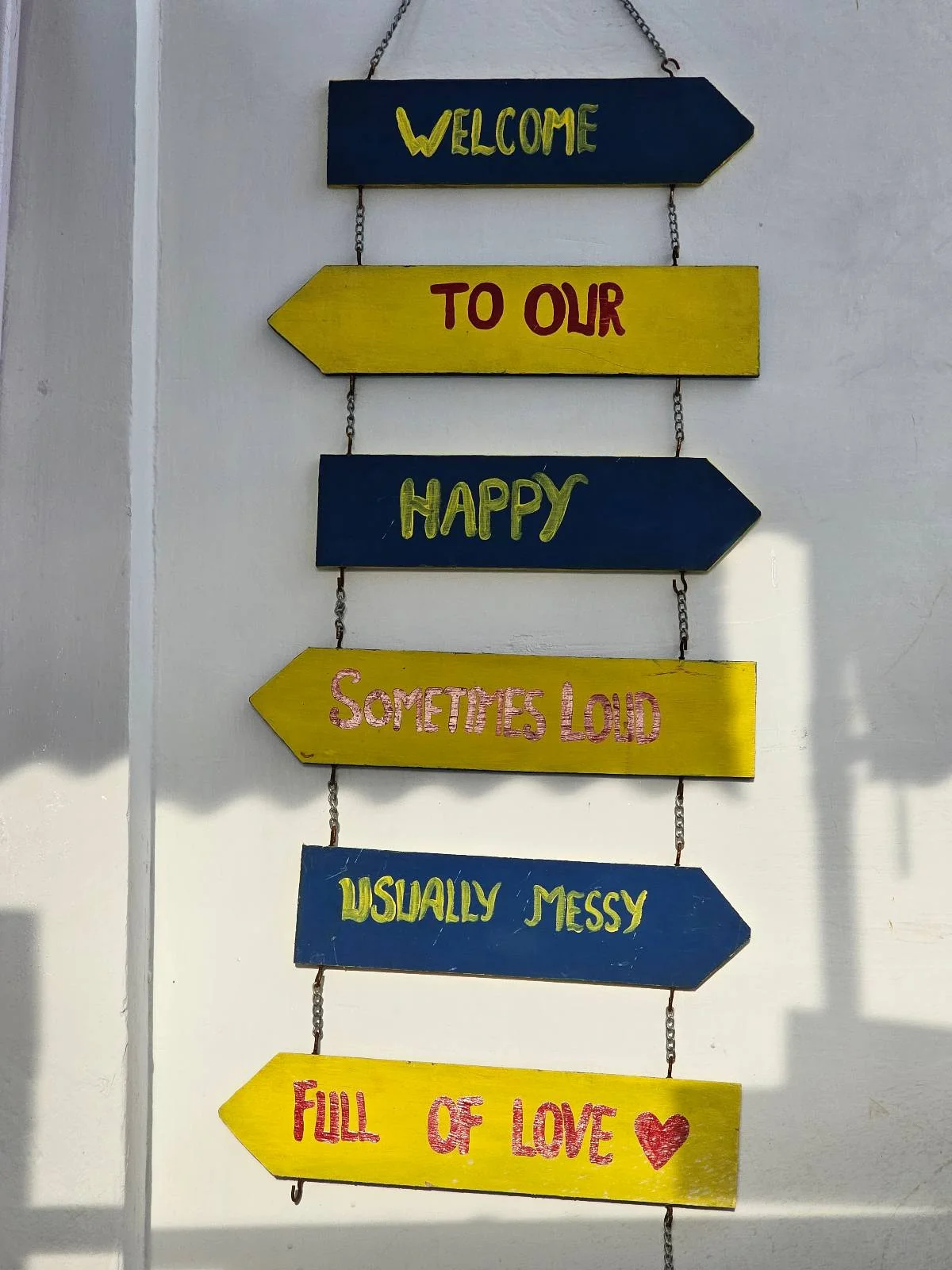 A wall decoration with six colorful arrow signs reads: "WELCOME," "TO OUR," "HAPPY," "CRAZY FUN HOME," "INSANELY MESSY," and "FULL OF LOVE" with a red heart at the end.