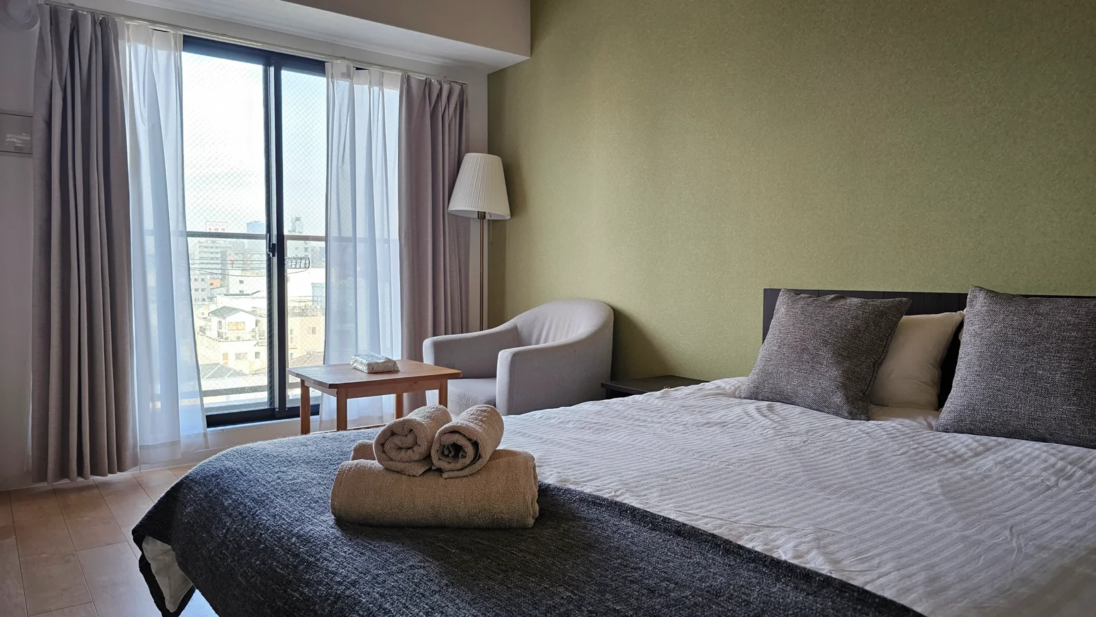 Modern bedroom with a neatly made bed, two gray pillows, rolled towels, a light gray armchair, a small table, a floor lamp, and a large window with sheer curtains letting in natural light.