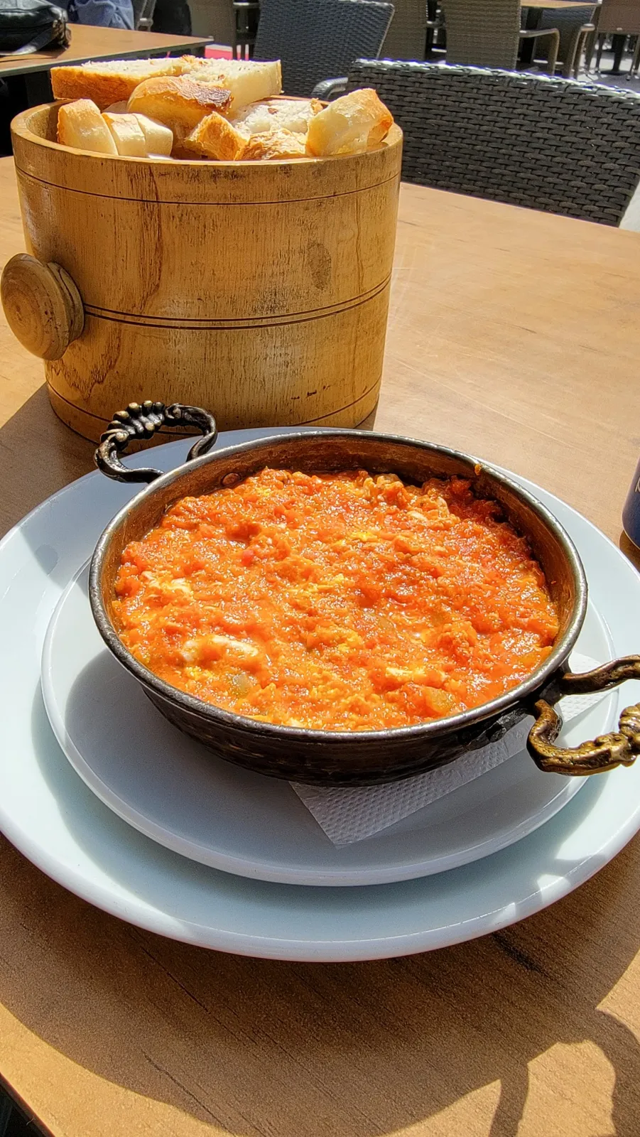 A skillet filled with a bright orange dish, possibly scrambled eggs or a stew, is placed on a plate. A wooden basket with sliced bread is in the background on a wooden table.