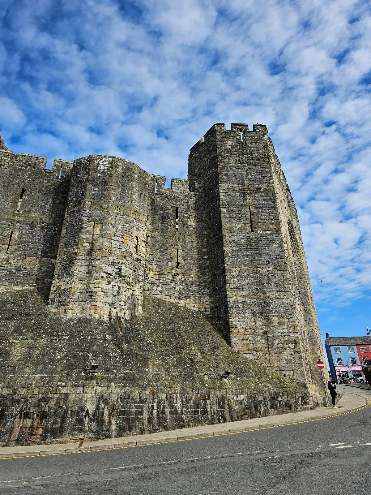 A tall, medieval stone castle wall with round towers stands under a blue sky filled with scattered clouds. A street and a colorful building are visible at the edge of the image.