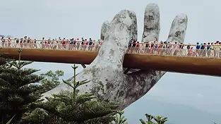 A wide shot of the Golden Bridge in Ba Na Hills, Da Nang, featuring a curved pedestrian walkway supported by two massive, weathered stone-like hands emerging from the lush green mountain.