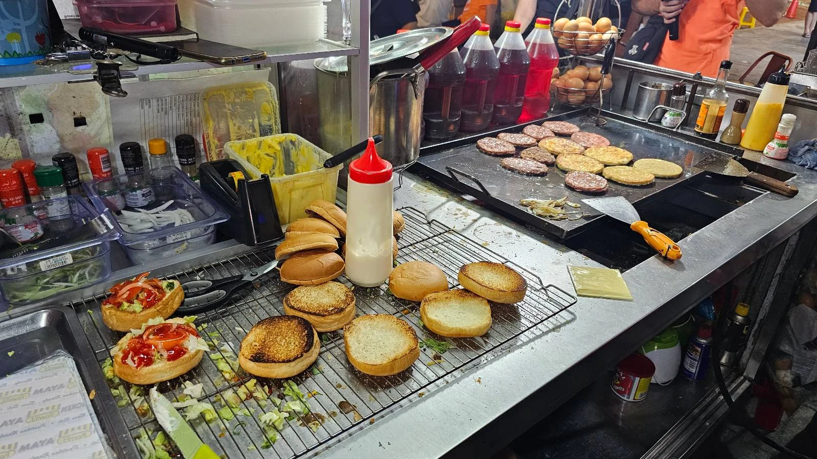 A street food stall with burger buns on a rack and patties cooking on a griddle. Condiments, sauces, and fresh vegetables are visible. Plastic bottles with red and yellow liquids are in the background.