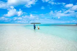 Two people standing in crystal-clear shallow lagoon water with white sand, turquoise sea, and a small tropical island under a bright blue sky.