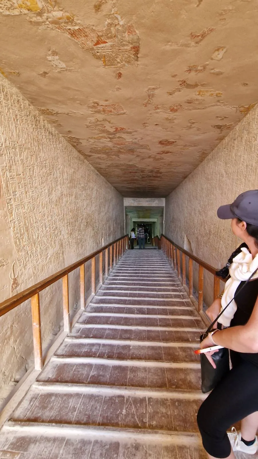 A person wearing a cap and black clothing descends a narrow, steep staircase with wooden railing, heading towards a dimly lit passage or room at the bottom.