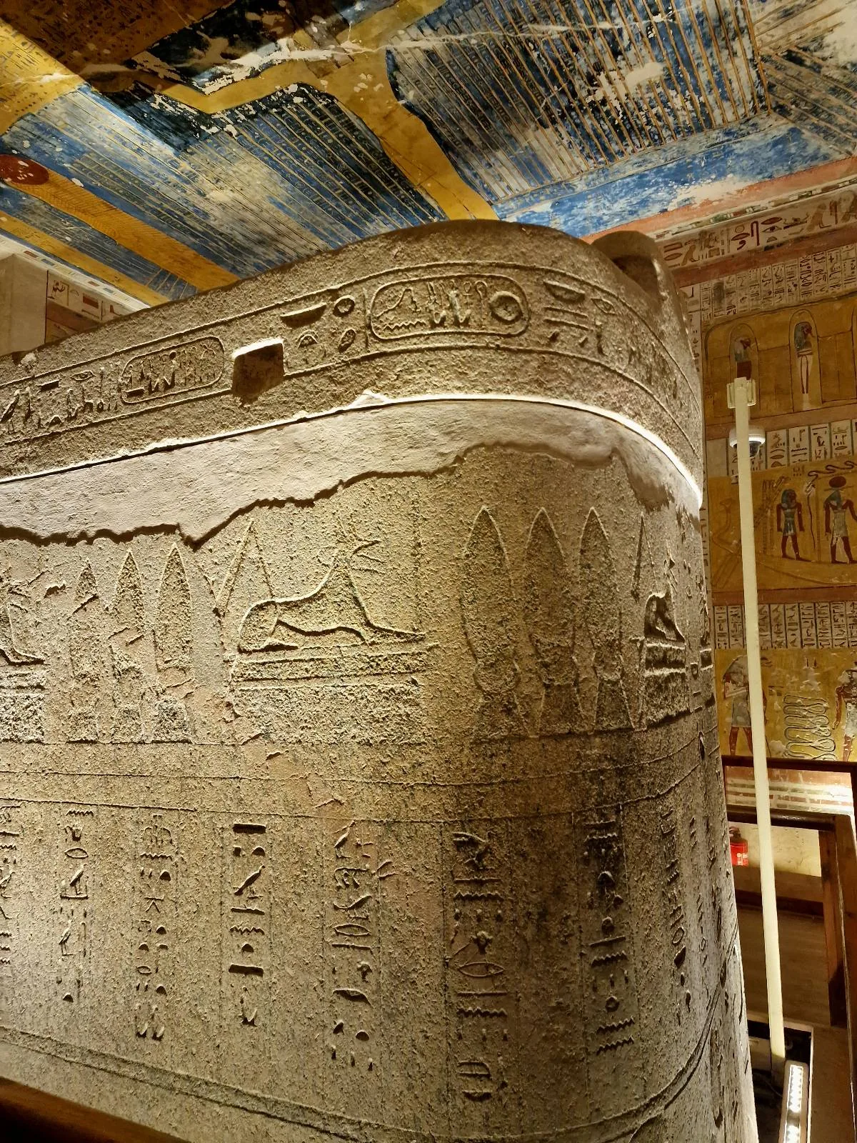 A sarcophagus with hieroglyphic inscriptions and carvings is in an ancient Egyptian tomb with detailed paintings on the walls and ceiling.