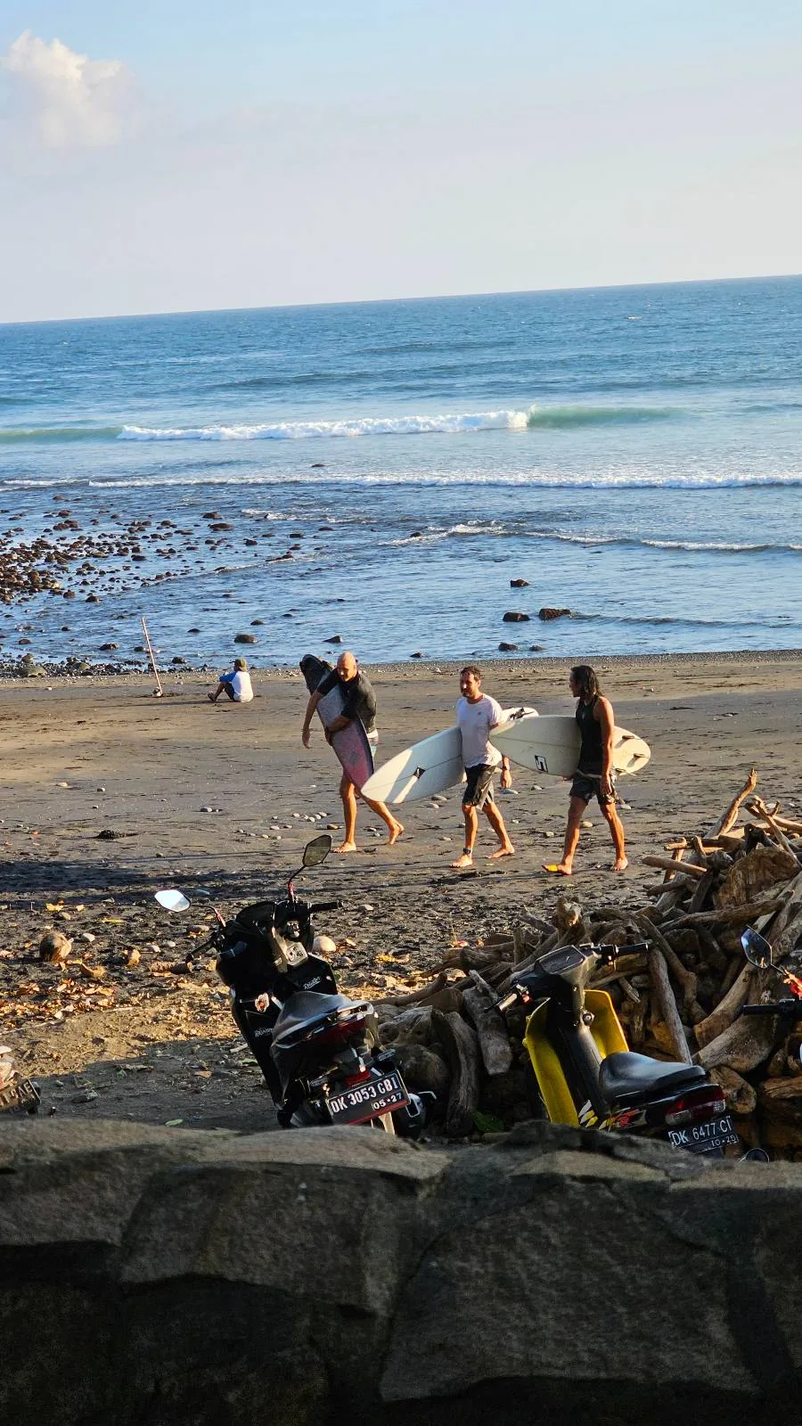 Three people with surfboards walk along a rocky beach towards the ocean. Another person sits near the shore. Two motorcycles are parked in the foreground. The sea is calm with gentle waves under a clear sky.